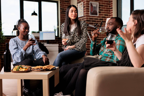 Happy young people enjoying drinking alcohol beverages together while celebrating birthday event. Diverse group of close friends relaxing together at home while hosting wine party in living room. - Photo, image