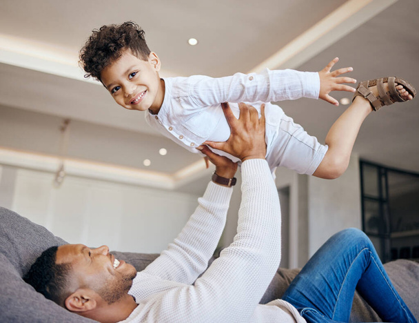 Joyful young dad lying on couch and lifting happy excited little boy in the air. Mixed race family having fun at home. Happy kid bonding with his father. - 写真・画像