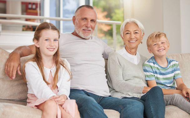 Two siblings relaxing with their grandparents together at home on a couch. Sister and brother visiting grandparents. A happy caucasian mature couple bonding with their adopted kids in the living room. - Foto, Imagen