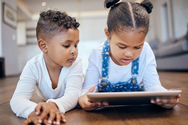 Δύο μικρά παιδιά μικτής φυλής χρησιμοποιώντας ένα ψηφιακό tablet, ενώ βρίσκονται μαζί στο πάτωμα στο σπίτι. Νεαρά αγόρια και κορίτσια αδέλφια περιήγηση στο διαδίκτυο για να παίξουν παιχνίδια και να μάθουν από εκπαιδευτικές εφαρμογές. - Φωτογραφία, εικόνα