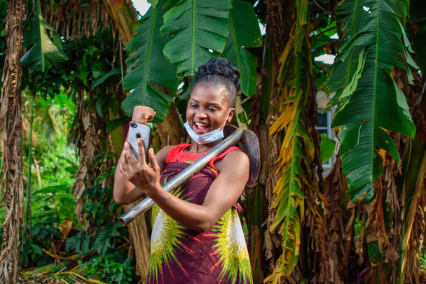 A female african farmer with nose mask and a farming hoe on her shoulder happily looks into a smart phone she's holding in a banana farm or plantation - Photo, Image