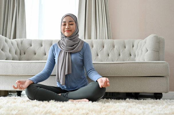 Peaceful millennial Asian Muslim woman with hijab practicing yoga, concentrated meditating, doing a lotus pose in the living room. - Fotoğraf, Görsel