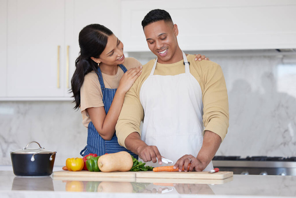 A young loving latin couple cooking a healthy dinner together at home. A loving man cutting carrots and other vegetable foods, preparing a meal and his girlfriend holds him and smiles in the kitchen. - Foto, imagen