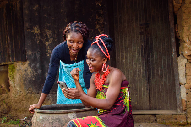 Two female Africans happily looking into the smart phone outside a village mud house that have a big water calabash - 写真・画像
