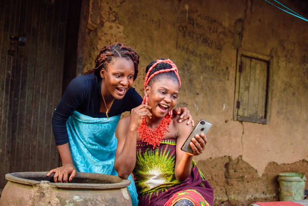 Δύο γυναίκες Αφρικάνες ψάχνουν με χαρά το έξυπνο τηλέφωνο έξω από ένα λασπωμένο σπίτι του χωριού που έχει μια μεγάλη νεροκολοκύθα - Φωτογραφία, εικόνα