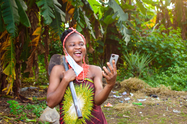 Samica afrykańskiego rolnika używająca maski nosowej, miejscowych koralików i motyki na ramieniu szczęśliwie patrzy na smartfon na farmie bananów lub plantacji bananów. - Zdjęcie, obraz