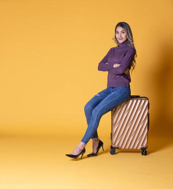 beautiful young woman with suitcase, sitting near chair, looking at camera, standing against blue background - Photo, image