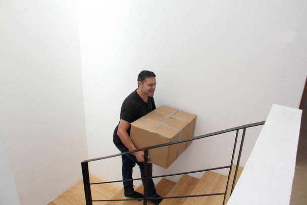 Latino adult man carries a heavy cardboard box up stairs which causes severe pain in his lower back and back - Фото, изображение