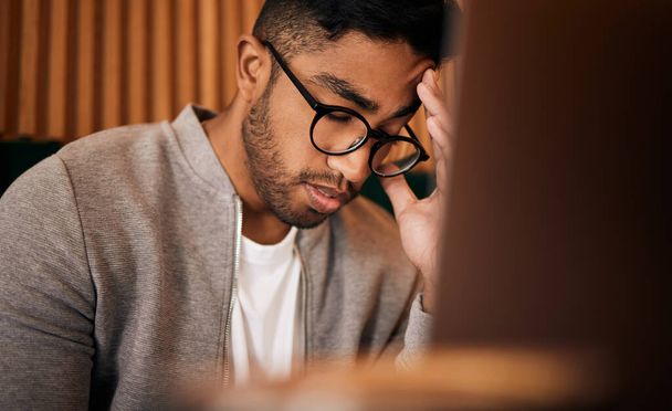 Young stressed indian man or student struggling with headache while browsing on a laptop in a cafe. One tired and frustrated guy suffering with burnout and pressure while studying or working freelance - Foto, imagen