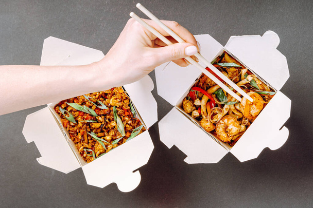 Wok boxes. Asian glass noodles with seafood and rice with vegetables. Female hand holding chopsticks and taking shrimp. - Foto, imagen