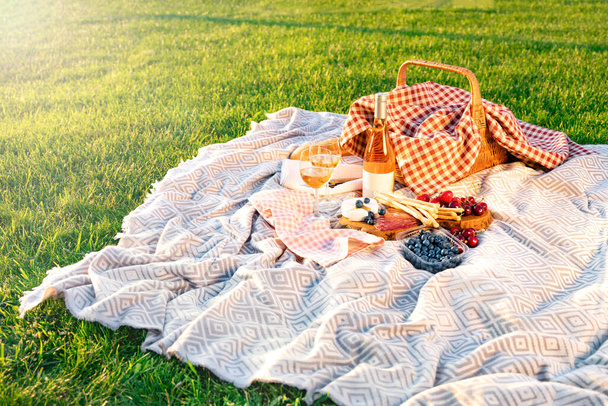 Picnic basket, wine, berries, jamon, cheese and bread are on the blanket. Picnic setting on a blanket in a meadow at sunset. - Фото, зображення