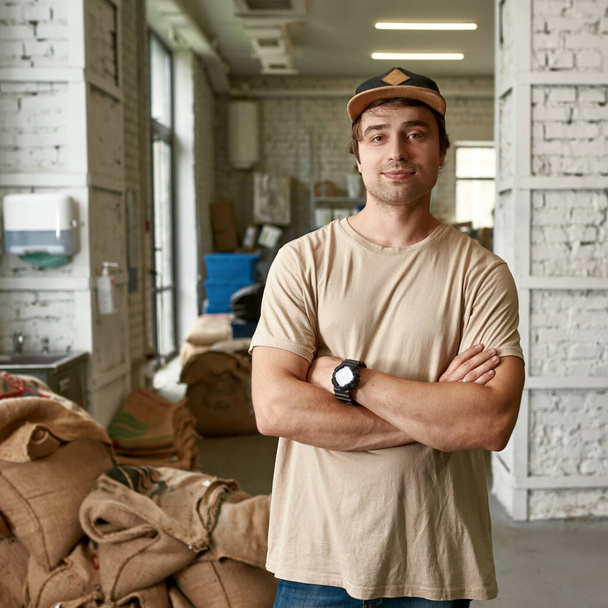 Young caucasian male barista or worker posing on coffee factory or warehouse. Coffee process making, production and storage. Small business. Man with crossed arms looking at camera - Valokuva, kuva