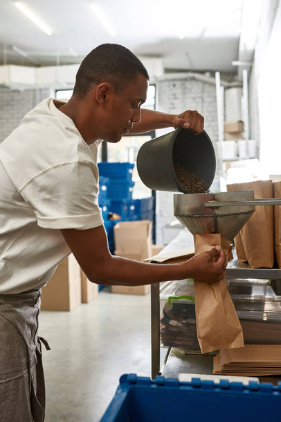 Male worker or barista pouring out roasted coffee beans from bucket in package on factory or coffee shop. Coffee process making and production. Small business. Young focused black man wearing apron - Foto, Imagen