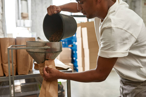 Male worker or business owner pouring out roasted coffee beans from bucket in package on factory. Coffee process making and production. Small business. Young focused black man wearing apron - Photo, Image