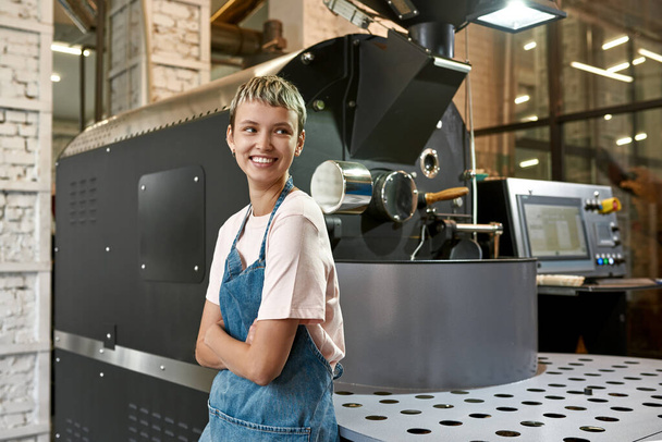 Young caucasian female worker or business owner near industrial coffee bean roasting machine on factory. Coffee process making and production. Small business. Modern automated manufacturing equipment - Foto, Bild