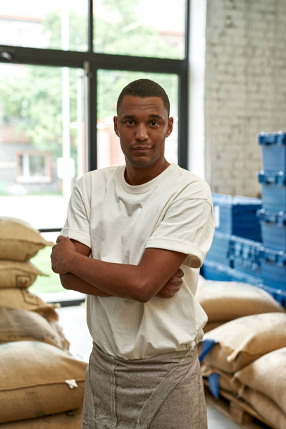 Front view of male worker near plastic baskets and sacks on coffee factory or warehouse. Coffee process making and production. Small business. Young black man with crossed arms looking at camera - Fotoğraf, Görsel