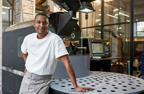 Young smiling black male worker or business owner near industrial coffee bean roasting machine on factory. Coffee making and production. Small business. Modern automated manufacturing equipment - Foto, Bild