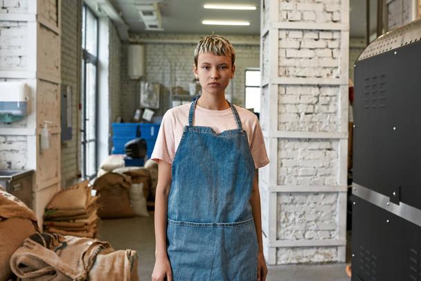 Young caucasian girl barista or worker looking at camera on coffee factory or warehouse. Coffee process making and production. Small business. Modern automated manufacturing equipment - 写真・画像