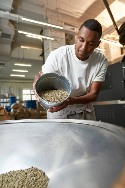Bottom view of young black male worker or barista pouring out green coffee beans with bucket in metal grinder. Coffee process making and production. Small business. Modern manufacturing equipment - Foto, Imagem