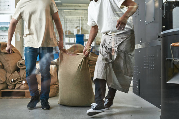 Cropped of multiethnic male workers carrying textile sack with coffee beans on factory or warehouse. Coffee process making and production. Small business. Teamwork. Black man wearing apron - Foto, Imagem