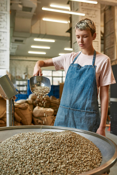 Young caucasian female worker pouring out fresh organic green coffee beans with iron scoop in metal grinder. Coffee process making and production. Small business. Modern manufacturing equipment - Fotografie, Obrázek