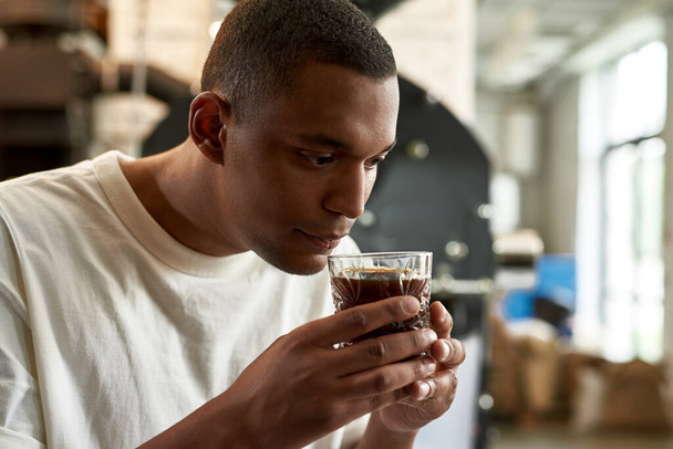 Professional male barista sniffing fresh aromatic natural coffee in glass on blurred factory. Young focused black man checking and studying coffee flavor. Coffee process making. Small business - Foto, Imagem