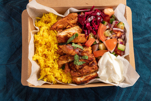 Традиционная восточно-средиземноморская еда из жареной курицы, желтого риса, свежего салата и йогурта в картонной упаковке - Фото, изображение