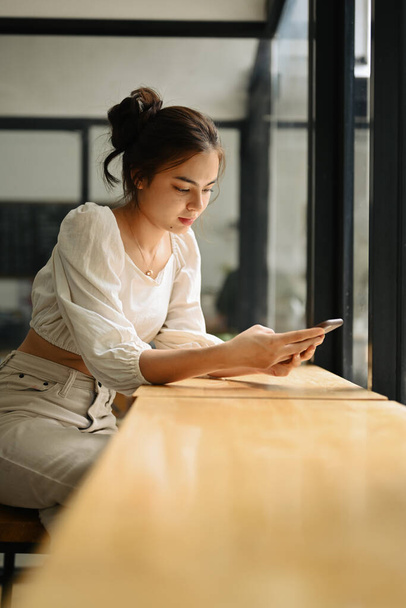 Urocza młoda kobieta siedzi przy oknie w kawiarni i wysyła wiadomości na smartfonie. - Zdjęcie, obraz