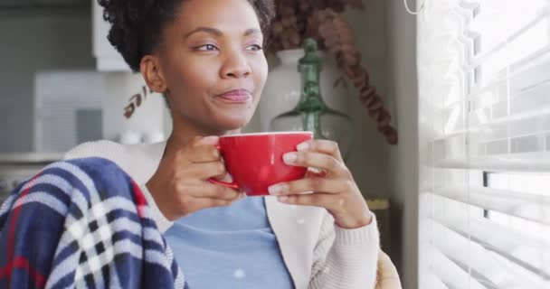 Video of thoughtful african american woman drinking coffee and looking outside window. Lifestyle, relax, and spending free time at home concept. - Materiał filmowy, wideo
