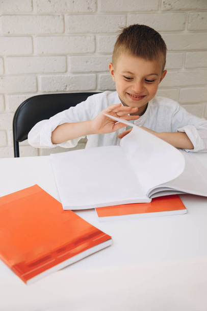 A boy in a white shirt at a white table flips through red textbooks and smiles. Back to school concept - Φωτογραφία, εικόνα