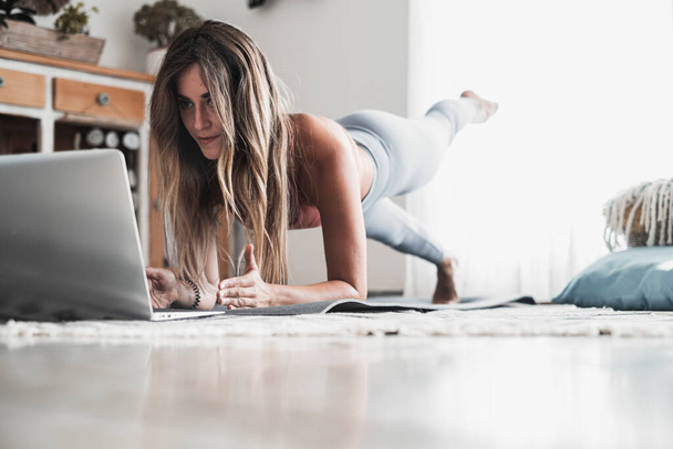 Κατάλληλο και υγιεινό τρόπο ζωής νεαρή γυναίκα κάνει σανίδα θέση στο σπίτι ψάχνει ένα μάθημα για το laptop. Αθλητισμός και ενεργή προπόνηση στο διαδίκτυο. Μετά το κανάλι στο διαδίκτυο για τη θετικότητα και την υγεία του σώματος - Φωτογραφία, εικόνα