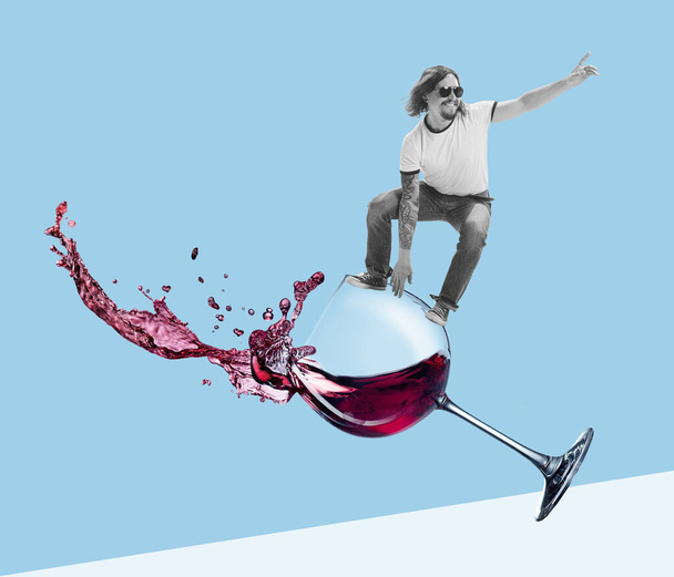 Contemporary art collage. Cheerful young man surfing on red wine glass isolated on blue background. Weekends. Concept of party, fun, retro design, creativity, imagination, inspiration, artwork and ad - Photo, Image