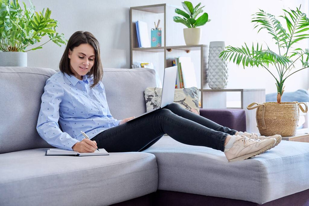 Young business woman working from home using laptop making notes sitting on sofa. Freelance, remote work, online business, technology, sales, e-learning, e-education concept - Fotó, kép