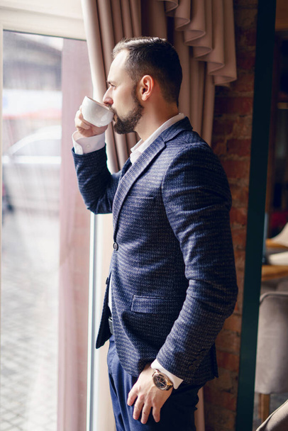 Ein junger Mann im Businessanzug trinkt am Fenster Kaffee. Ein junger Mann mit Bart in einem Café. - Foto, Bild