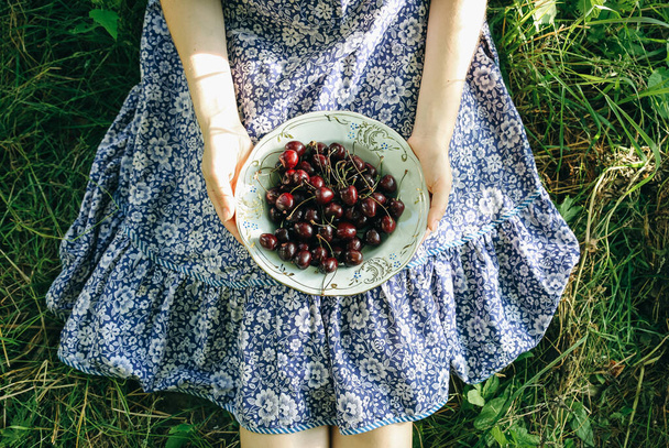 Ragazza è seduta sull'erba in abito vintage blu. Quella donna tiene le ciliegie. Rustico di frutta estiva posa piatta. Sano vegetariano concetto di stile di vita alimentare ecologico. - Foto, immagini