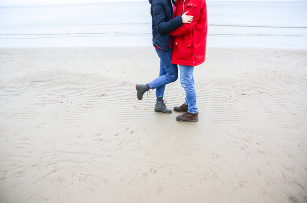 Zwei Hipster stehen am kalten Strand. Paar umarmt und hält Händchen. Liebesgeschichte in der Nähe des Ozeans. Wintersaison auf dem Meer. Stilvolle Stiefel im Sand. Mann in roter Jacke. - Foto, Bild