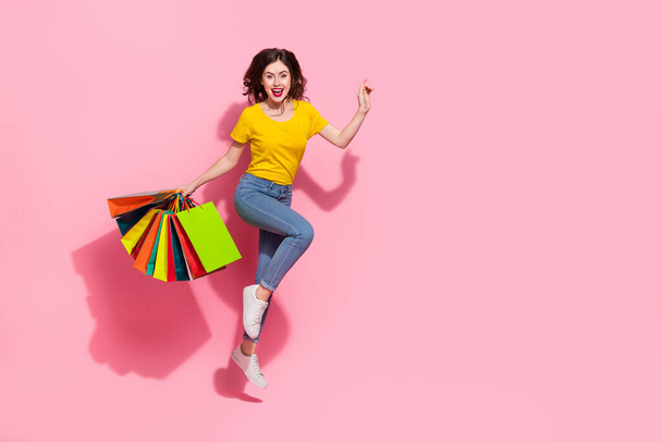 Full size photo of impressed young brunette lady jump with bags wear t-shirt jeans sneakers isolated on pink background. - Foto, imagen
