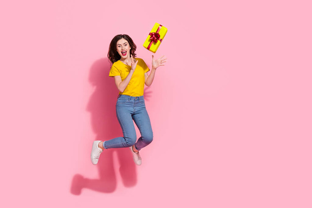 Full length photo of impressed young brunette lady jump with present wear t-shirt jeans sneakers isolated on pink background. - 写真・画像