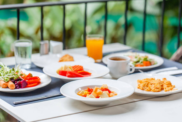 Buffet breakfast on terrace in outdoors restaurant at tropical hotel or resort. Brunch table with food and drinks, fried eggs, coffee cup, fruits. Summer holidays, vacations, tourism, travel concept. - Фото, изображение