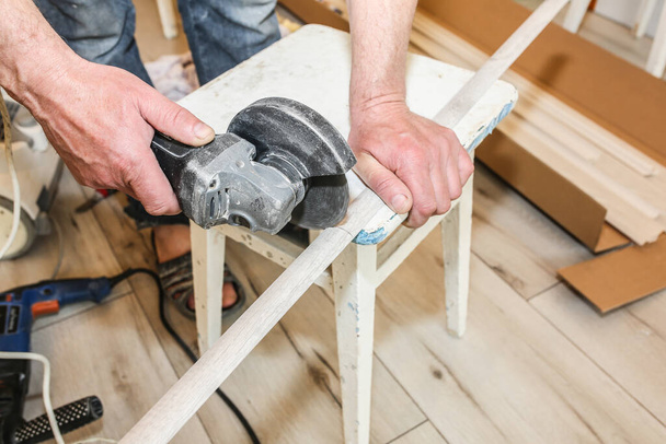 Man is cutting baseboard. Repair works indoors. Renovation in the flat.  Angle grinder in hands. - Foto, imagen