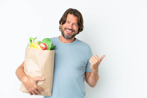 Senior dutch man holding grocery shopping bag over isolated background pointing to the side to present a product - Foto, Bild