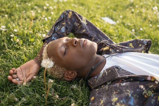 Portrait of young African American guy with stylish hair laying on grass, relaxing in park. Fashion model posing for pictures. Beauty concept - 写真・画像
