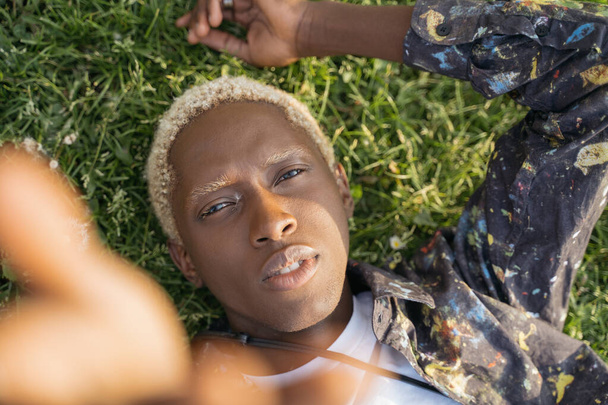 Портрет молодого афроамериканца со стильными волосами, делающего селфи, смотрящего в камеру, лежащую на траве. Мода модель позирует для фотографий на открытом воздухе. Концепция красоты - Фото, изображение