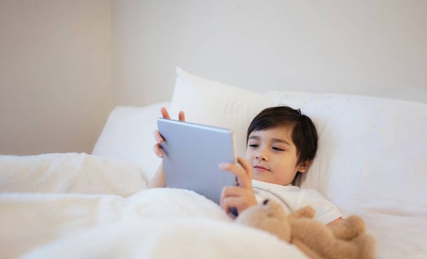 Счастливый малыш, лежащий в кровати, держа в руках стол, смотрящий мультики и болтающий с друзьями на цифровом планшете, Симпатичный мальчик, играющий в игры онлайн в Интернете, Ребенок, расслабляющийся утром перед тем, как пойти в школу - Фото, изображение