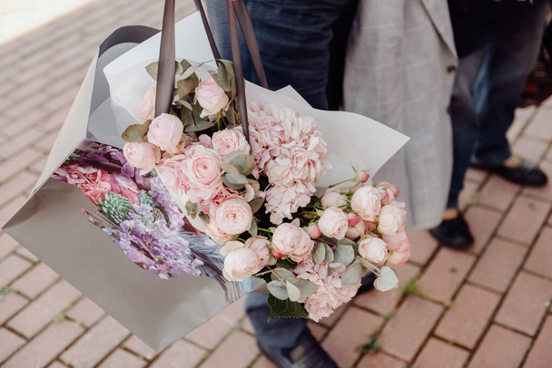 A bouquet of pink roses lies in a craft bag with fabric ribbons handles in hands against the background of a tiled floor - Фото, изображение
