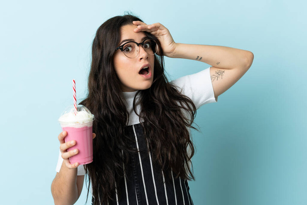 Young woman with strawberry milkshake isolated on blue background doing surprise gesture while looking to the side - Photo, Image