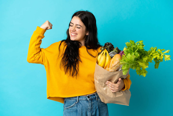 Young woman holding a grocery shopping bag isolated on blue background doing strong gesture - Photo, Image