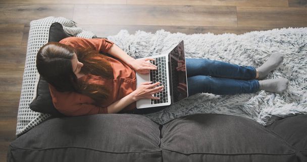 Genç kadın rahat bir kanepede uzanıyor ve dizüstü bilgisayarda yazıyor. Üst düzey görüş, evde çalışma, rahat ofis, uzak çalışma, blog, yazar, yazışma, karantina, serbest çalışma. Yüksek kalite 4k görüntü - Fotoğraf, Görsel