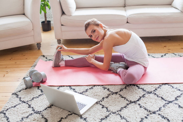 Pretty fit woman wearing sportswear doing fitness stretching exercises and using laptop at home. Sport and workout concept. - Zdjęcie, obraz