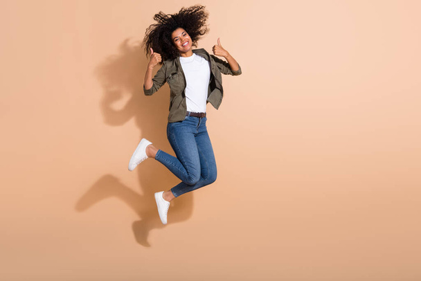 Full length body size view of attractive cheerful girl jumping showing thumbup advert isolated over beige pastel color background. - Foto, Imagem